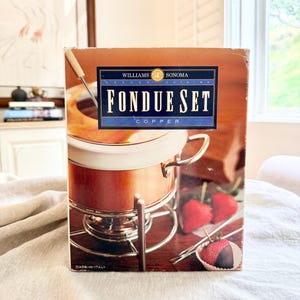 May include: A Williams Sonoma copper fondue set in its original box. The box features a photo of the fondue pot, strawberries, and the words "Fondue Set Copper". The set includes a copper pot and a silver stand.