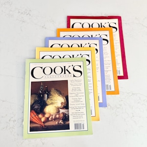 May include: A stack of Cook's Illustrated magazines with colorful covers. The magazines feature the title "Cook's Illustrated" in a bold font. The covers are in shades of green, yellow, blue, orange, and red. The front cover of the magazine in the foreground features a still life of vegetables.
