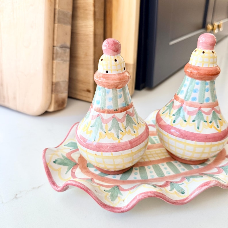Pu&ograve; includere: Una coppia di saliera e pepiera in ceramica con vassoio coordinato dai bordi ondulati, caratterizzata da un motivo dipinto a mano a righe, quadretti e fiori nei toni del rosa, giallo e verde su fondo bianco, con forma arrotondata.