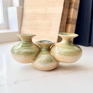 May include: A cluster of three small, handcrafted ceramic vases. Each vase has a rounded body and a flared opening. The vases are glazed in a gradient of colors, transitioning from a creamy beige at the base to a soft green at the top.