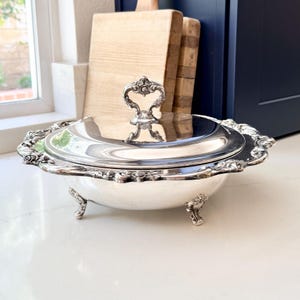 May include: A silver-plated serving dish with a lid, ornate handle, and decorative rim. The dish has three small feet and is set on a white surface. The background includes wooden cutting boards and a window.