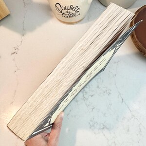 May include: A book with aged, off-white pages and a decorative black and white cover. The book is held open, revealing the interior pages. The background includes a white countertop and ceramic kitchenware.