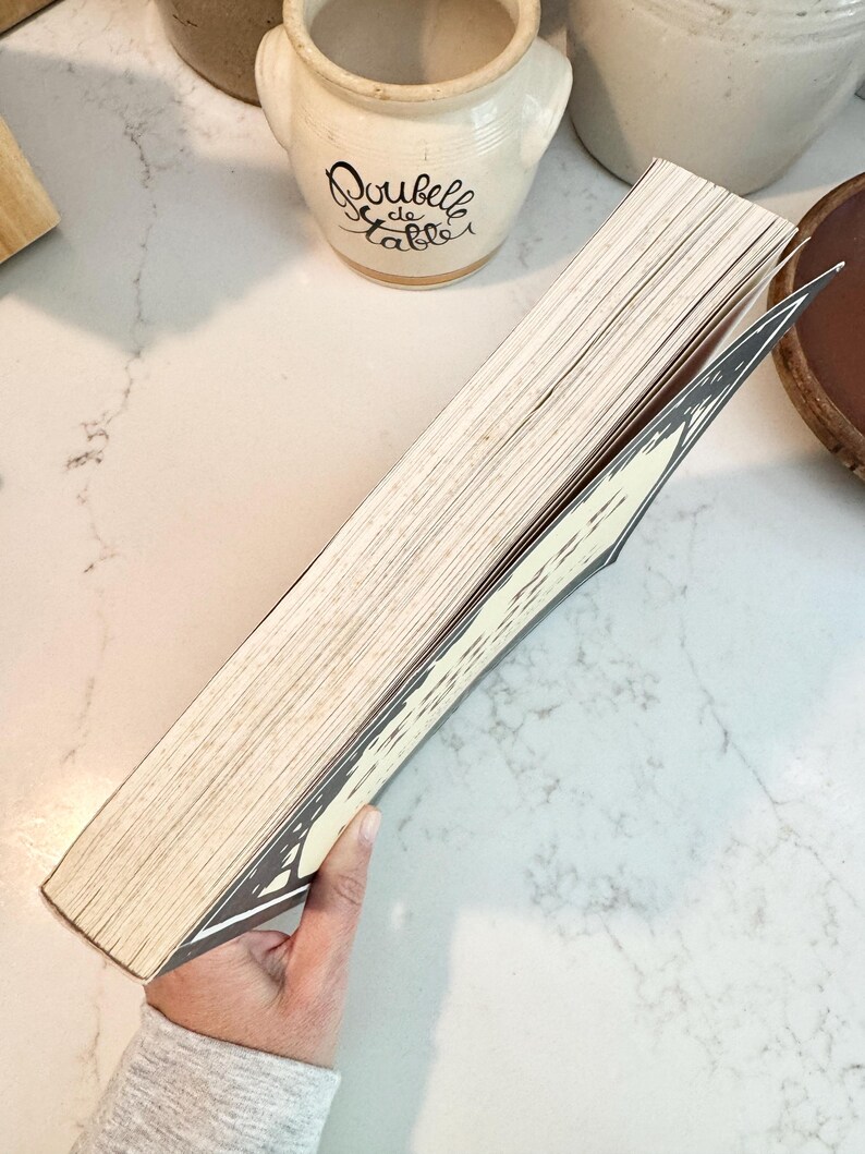 May include: A book with aged, off-white pages and a decorative black and white cover. The book is held open, revealing the interior pages. The background includes a white countertop and ceramic kitchenware.