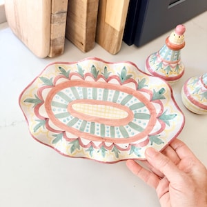 Pu&ograve; includere: Un piatto ovale in ceramica con bordo smerlato, caratterizzato da un motivo dipinto a mano con strisce verde salvia, bordi rosa e un design a griglia giallo centrale. Il piatto ha una base color crema con motivi decorativi a foglia verde.