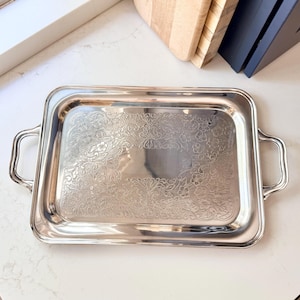 May include: A rectangular silver-toned metal serving tray with two curved handles on the sides. The center of the tray features an intricate, embossed floral and scrollwork pattern, while the edges remain smooth and polished with a reflective finish.