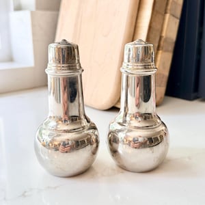 Può includere: Un paio di saliere e pepiere d'argento vintage. Le saliere e le pepiere hanno una base bulbosa, un collo sottile e un tappo con piccoli fori. La superficie riflettente mostra un riflesso distorto. Sono su una superficie bianca.