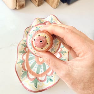 Pu&ograve; includere: Un salino o pepaiolo in ceramica con parte superiore arrotondata e vassoio smerlato abbinato. Il set presenta un motivo dipinto a mano nei toni del terracotta, verde salvia e giallo, con un foro centrale sul coperchio per l'uso.