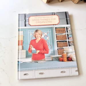 May include: A hardcover book titled Good Things for Organizing by Martha Stewart Living. The cover features a person in a red sweater working at a desk with storage baskets, clear bins, and yarn balls visible in a light blue craft room setting.