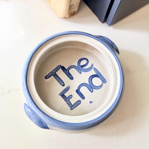 May include: A round, ceramic bowl with a white interior and a blue rim. The words "The End" are hand-painted in blue inside the bowl. The bowl has two small handles and is set on a white surface.