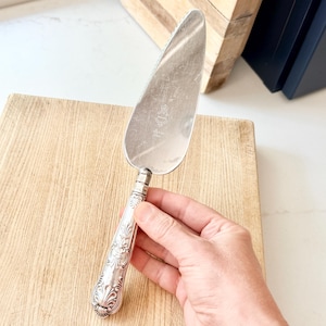May include: Silver cake server with a decorative handle featuring floral and scroll designs. The server has a wide, triangular blade with a serrated edge. The handle is held by a person.