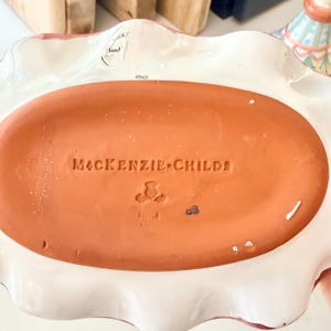 Pu&ograve; includere: Il lato inferiore di un piatto ovale in ceramica con una base color terracotta. Il testo MACKENZIE-CHILDS &egrave; impresso al centro, accompagnato da un piccolo timbro a forma di cardo e dalle lettere SC. Un bordo bianco ondulato circonda la base.