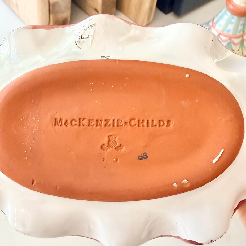 Pu&ograve; includere: Il lato inferiore di un piatto ovale in ceramica con una base color terracotta. Il testo MACKENZIE-CHILDS &egrave; impresso al centro, accompagnato da un piccolo timbro a forma di cardo e dalle lettere SC. Un bordo bianco ondulato circonda la base.