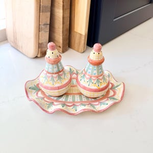 Pu&ograve; includere: Una coppia di saliera e pepiera in ceramica con vassoio ovale abbinato, caratterizzata da un motivo dipinto a mano con forme geometriche, strisce e punti rosa, verdi e gialli su base color crema con bordi smerlati.