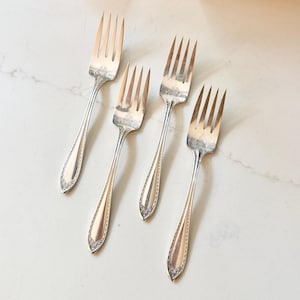 May include: Four silver forks with tines and ornate handles are arranged on a white marble surface. The forks have a decorative pattern near the tines and along the handle edges, suggesting a vintage or antique style. The forks are angled towards the center.