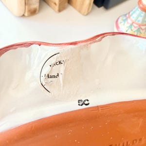 Pu&ograve; includere: Una ciotola in ceramica con bordo ondulato dipinto di rosso e interno bianco. Un adesivo bianco circolare parzialmente strappato &egrave; attaccato alla parete interna, con il testo "Hand" e "CK". Le lettere "SC" sono stampate in inchiostro nero.