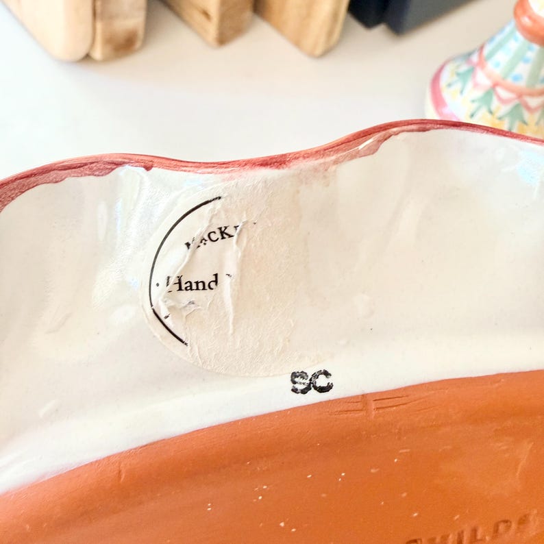 Pu&ograve; includere: Una ciotola in ceramica con bordo ondulato dipinto di rosso e interno bianco. Un adesivo bianco circolare parzialmente strappato &egrave; attaccato alla parete interna, con il testo "Hand" e "CK". Le lettere "SC" sono stampate in inchiostro nero.