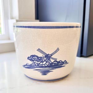 May include: A cream-colored ceramic planter featuring a blue horizontal band around the top rim and a blue illustration of a windmill in a landscape on the front. The surface of the planter has a visible crackle glaze finish throughout the exterior.