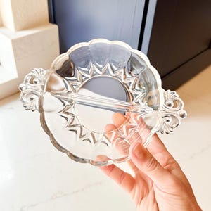 May include: Clear glass serving dish with a scalloped edge and two ornate handles. The dish is divided into two sections and features a starburst design in the center. A decorative piece for serving snacks or appetizers.
