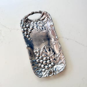 May include: A rectangular silver-toned metal tray featuring an embossed design of grape clusters and vine leaves. The tray has a smooth, reflective central surface and a handle integrated into the top edge, with detailed relief work throughout.