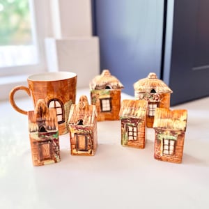 Op de afbeelding: Een verzameling keramische miniatuurhuisjes en een mok met een boomschorsontwerp. De huisjes hebben rieten daken, ramen en deuren. De mok heeft een handvat en een vergelijkbare boomschorstextuur. Het kleurenpalet omvat bruin- en beigetinten.