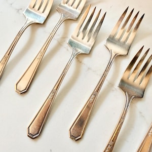 May include: Close-up of several silver forks arranged diagonally. The forks have a classic design with tines and long handles. The handles feature a decorative floral pattern. The forks are set against a white marble surface.