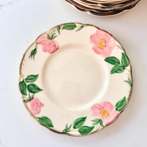 May include: Cream-coloured ceramic plate with a scalloped edge, decorated with pink rose blossoms and green leaves. The plate has a gold-coloured rim and is part of a set. A few plates are stacked in the background.