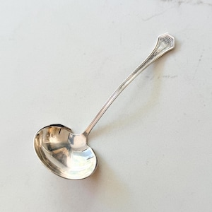 May include: A silver-toned metal ladle featuring a polished, rounded bowl and a long, slender handle. The handle terminates in a decorative, pointed end with intricate engraved detailing, providing a classic look for serving liquids or sauces.