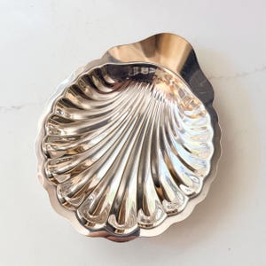 May include: A polished silver-toned metal dish shaped like a scallop shell. The dish features a series of deep, radiating grooves that extend from the hinge area to the scalloped outer edge, creating a reflective and textured surface throughout.