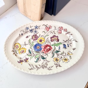 May include: Oval serving platter with a cream-colored background and a floral design. The flowers are in shades of red, yellow, blue, and purple, with green leaves. The rim has a rope-like texture.