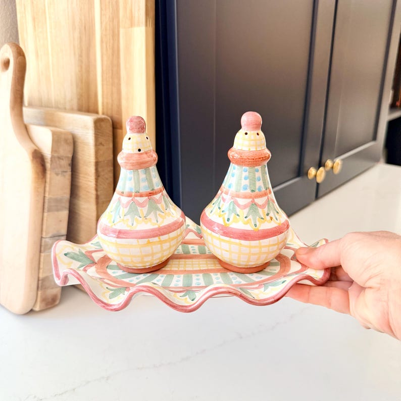 Pu&ograve; includere: Una coppia di saliera e pepiera in ceramica con vassoio abbinato dal bordo ondulato, caratterizzata da un motivo dipinto a mano a righe, quadri e disegni floreali nei toni del rosa, giallo e verde su base color crema lucida.