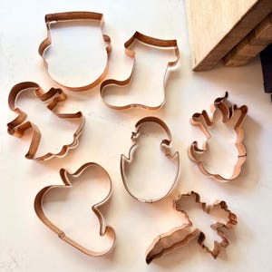 Può includere: Una collezione di formine per biscotti color rame in varie forme, tra cui una calza, un angelo, un cuore e un pulcino che esce da un uovo. Le formine sono disposte su una superficie chiara, suggerendo un tema di pasticceria.