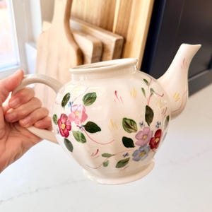 May include: A cream-colored ceramic teapot featuring a hand-painted floral pattern with red, pink, purple, and blue flowers, green leaves, and thin pink and yellow decorative lines. The teapot has a curved handle, a spout, and a fluted body design.