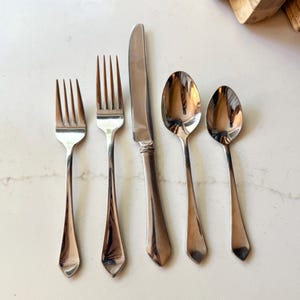 Könnte beinhalten: Ein Set aus fünf Edelstahl-Besteckteilen: zwei Gabeln, ein Messer und zwei Löffel. Das Besteck hat eine glänzende, reflektierende Oberfläche und ein klassisches Design. Das Besteck ist auf einer weißen Oberfläche angeordnet.