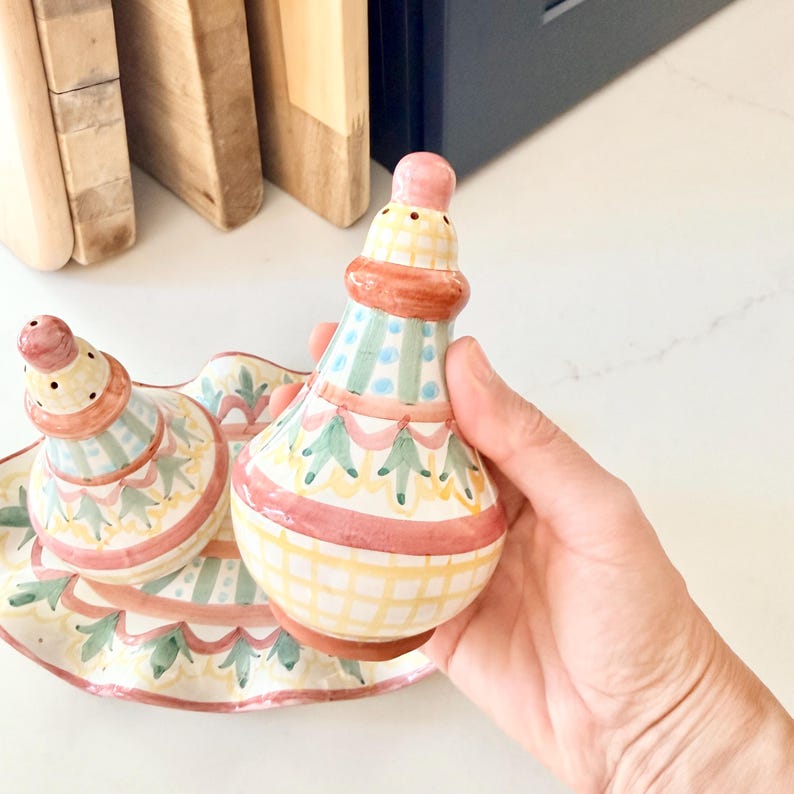 Pu&ograve; includere: Una coppia di saliera e pepiera in ceramica con vassoio ovale abbinato, caratterizzata da un motivo dipinto a mano di forme geometriche, strisce e disegni floreali rosa, gialli, verdi e blu su fondo color crema con finitura lucida.