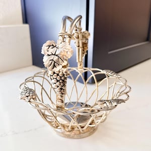 May include: A silver-toned metal basket featuring a lattice-style wire frame and a swing handle. The handle and the rim of the basket are decorated with detailed, raised relief motifs of grape clusters and vine leaves in a metallic finish.