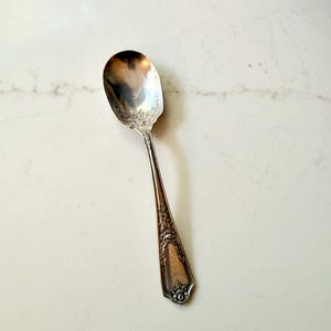 May include: A silver spoon with an oval bowl and a long, ornate handle. The spoon has a floral design on the bowl and handle. The spoon is set against a white marble background. The spoon is likely vintage or antique.