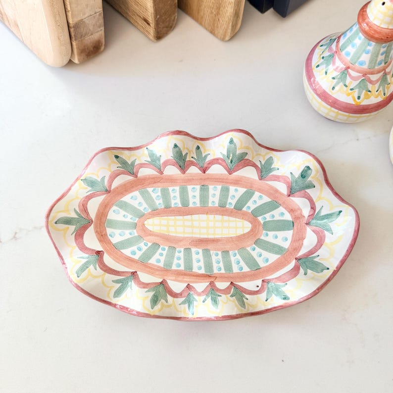 Pu&ograve; includere: Un piatto ovale in ceramica con bordo smerlato, caratterizzato da un motivo dipinto a mano con strisce verdi e gialle, una griglia centrale gialla e un bordo rosa con accenti a forma di foglia verde su una base color crema.