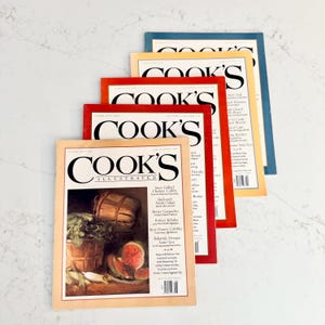 May include: A stack of Cook's Illustrated magazines with colorful covers. The magazines feature the title "Cook's Illustrated" in large black letters. The covers have images of food and recipes.