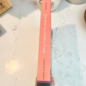 May include: A cookbook titled "The New California Cook" by Chronicle Books. The book's spine is a salmon color with white text. The bottom of the spine is dark with a white graphic. The book is held by a person.
