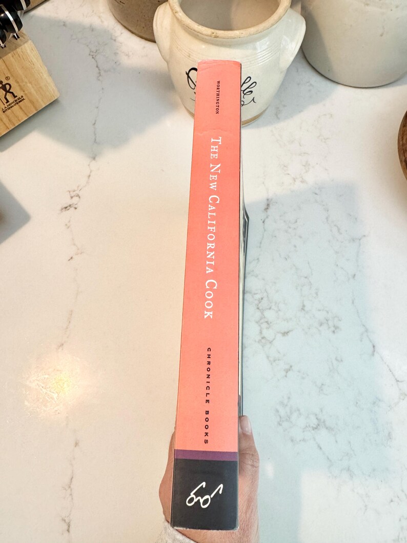May include: A cookbook titled "The New California Cook" by Chronicle Books. The book's spine is a salmon color with white text. The bottom of the spine is dark with a white graphic. The book is held by a person.