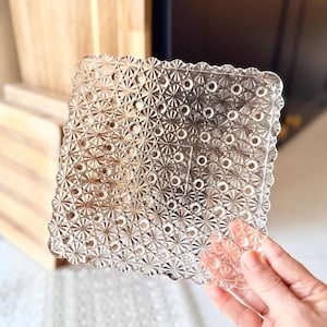 May include: A square, clear glass plate featuring a pressed, geometric pattern of alternating starburst motifs and small circular indentations. The edges of the plate are scalloped, creating a decorative border around the textured surface.