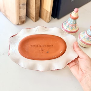 Pu&ograve; includere: Un piatto ovale in ceramica con un bordo bianco arricciato e una base color terracotta che presenta il testo in rilievo MacKenzie-Childs. La parte inferiore del piatto &egrave; liscia e piatta, in contrasto con il bordo bianco decorativo e ondulato.