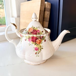 May include: White ceramic teapot with a floral rose pattern in shades of red, pink, and orange. The teapot has a curved spout, a gold-accented handle, and a matching lid with a finial. The teapot is likely for serving tea.