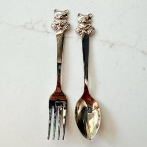 May include: A silver-colored fork and spoon set. Each utensil features a detailed teddy bear figure at the top of the handle. The fork has three tines, and the spoon has a rounded bowl. The set is designed for children.