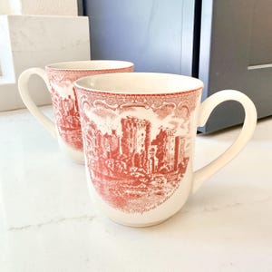 May include: A white ceramic mug with a black handle and the text "But First, Coffee" in a bold, black font. The mug is set against a blurred background, suggesting a cozy setting.