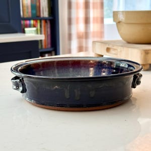 May include: A handmade, ceramic baking dish with a dark blue glaze. The dish has a round shape with two small handles and a reddish-brown base. The interior features a blend of blue and burgundy hues.