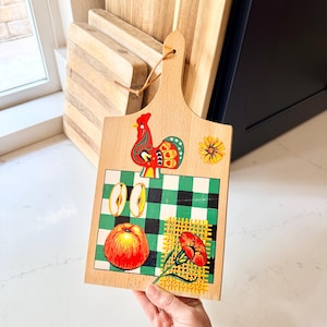 May include: A light-colored wooden cutting board with a handle and a leather hanging loop. The board features a colorful illustration of a red rooster, a yellow flower, two apple slices, a whole red apple, and a red flower on a green checkered background.