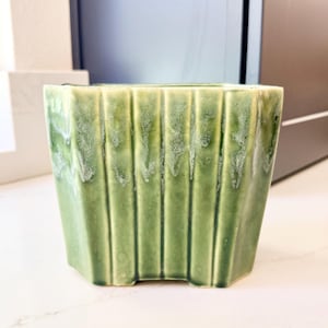 May include: A square, green ceramic planter with vertical ribbed detailing. The planter has a slightly distressed, glazed finish with a light green color. The planter is designed for indoor or outdoor use.
