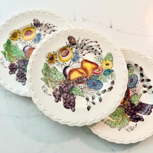 May include: Three white ceramic plates with a decorative fruit and floral design. Each plate features a border of woven texture. The center of each plate displays a colorful arrangement of grapes, pears, and flowers in shades of yellow, purple, and green.