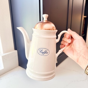 May include: A white ceramic coffee pot featuring a curved handle, a long spout, and a metallic copper-colored lid topped with a small white knob. The front of the pot displays the word Coffee in blue cursive script inside an oval border.